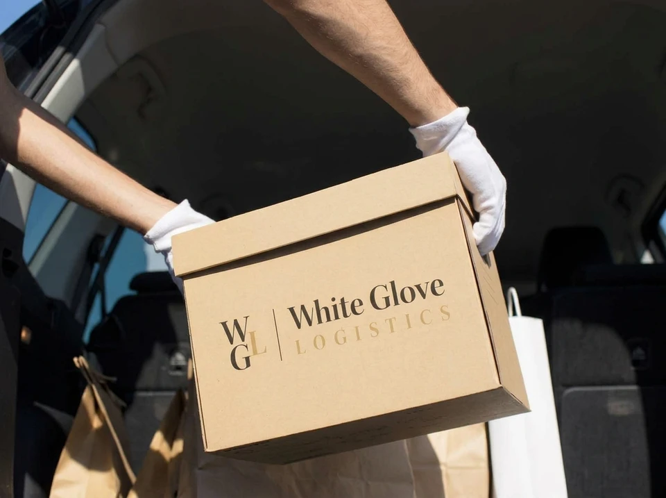 Déménageur en uniforme noir "White Glove Logistics", gants blancs, manipule un fauteuil Eames lounge chair en palissandre et cuir noir dans un salon luxueux